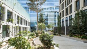 Cour urbaine moderne, agrémentée d'une végétation luxuriante, entourée de bâtiments contemporains en verre et en béton. L'atmosphère y est paisible et accueillante.