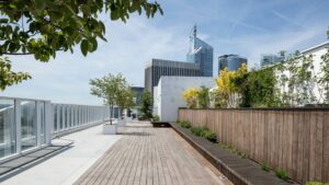 Jardin sur le toit avec terrasse en bois, jardinières modernes et végétation luxuriante. Garde-corps en verre à gauche, gratte-ciel du quartier de La Défense en arrière-plan sous un ciel bleu azur.