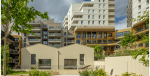 Complexe d'appartements moderne à l'architecture géométrique, avec ses façades beiges et ses grandes fenêtres. Des arbres et des plantes vertes agrémentent le cadre urbain.