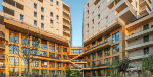 Un complexe d'appartements moderne avec de grandes baies vitrées et des façades beiges formant un V. Le ciel, clair et bleu, met en valeur le design épuré du bâtiment.