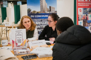 Deux femmes sont assises à une table lors d’un événement professionnel, discutant avec un homme. Des brochures et une pancarte indiquant « Kaufman & Broad » sont visibles. L’atmosphère est professionnelle.