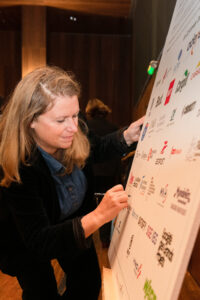 Une femme signe une grande affiche remplie de divers logos dans un cadre chaleureusement éclairé. Elle semble concentrée et engagée, transmettant un sentiment d’importance.